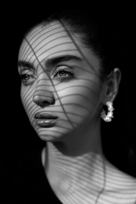 A black-and-white close-up portrait of a young woman wearing a pearl hoop earring, her face dramatically lit with striped shadows creating a striking chiaroscuro effect.