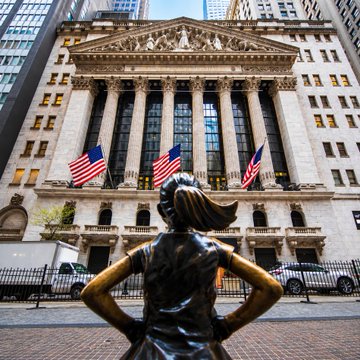 The "Fearless Girl" statue stands defiantly with hands on hips in front of the New York Stock Exchange, its grand columns and American flags framing the scene. The image captures the symbolic intersection of finance and emerging markets, tying directly to NYSE's new partnership with Blockchain.com to offer tokenized US stocks to millions of crypto users.