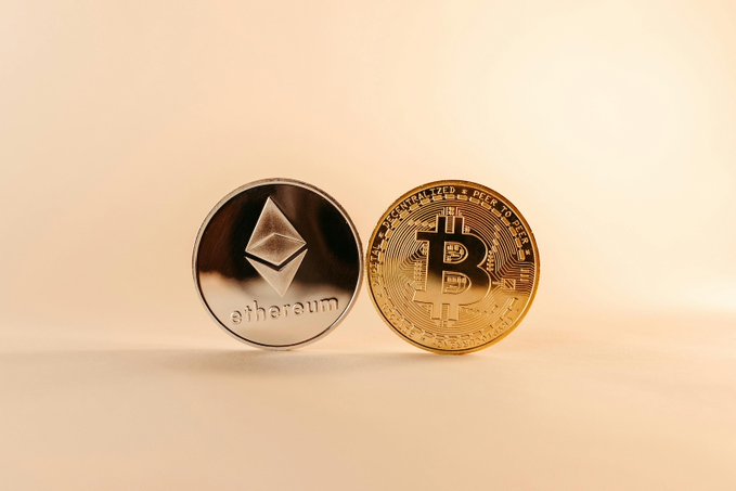 Two cryptocurrency coins—an Ethereum coin featuring its diamond logo and "ethereum" text, alongside a gold Bitcoin coin marked "BITCOIN DIGITAL · DECENTRALIZED · PEER TO PEER"—stand upright against a warm beige background, visually representing the two major digital assets driving the record-breaking $1.1B in combined weekly spot ETF inflows mentioned in the post.