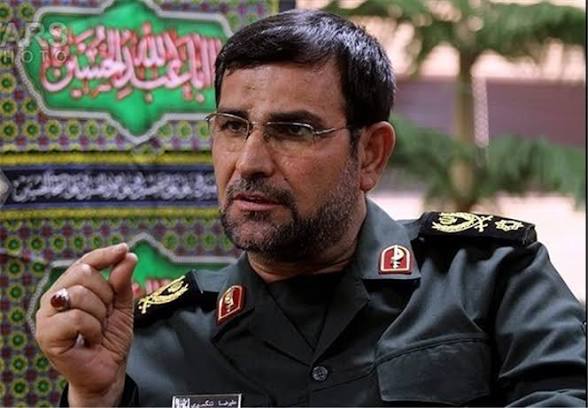 ارزبار بالعلم, a Persian military officer, speaks with a serious expression against a green and yellow background.