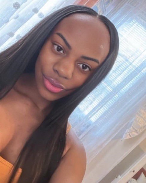 A young woman with long straight black hair takes a selfie indoors, wearing an orange top with sheer curtains visible in the background.