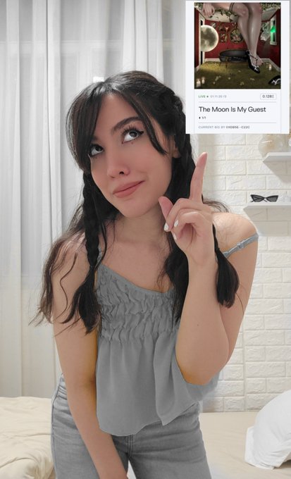 A young woman with dark braided hair in a grey off-shoulder top points to a Twitter/X post titled "The Moon Is My Quest" showing anime artwork of legs.