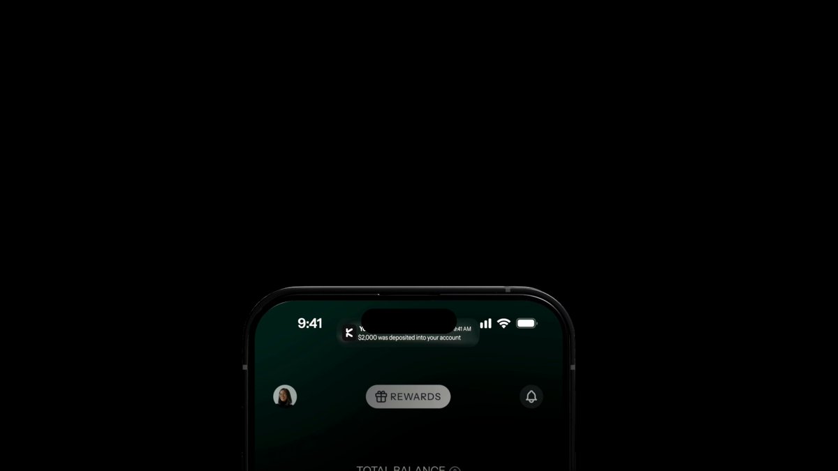 The image shows a cell phone screen displaying a message that reads "Watching @KASTxyz evolve lately feels a bit different. At first, it was just another crypto card project to many people. But little by little, it's becoming something much bigger". The phone is placed on a table with a brown background and the text is written in white.