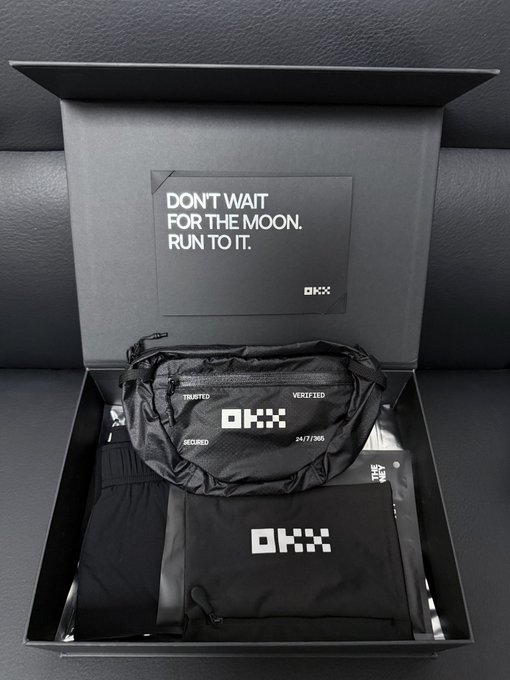 An open black OKX gift box containing athletic wear and a fanny pack, featuring a card that reads 'DON'T WAIT FOR THE MOON. RUN TO IT.'