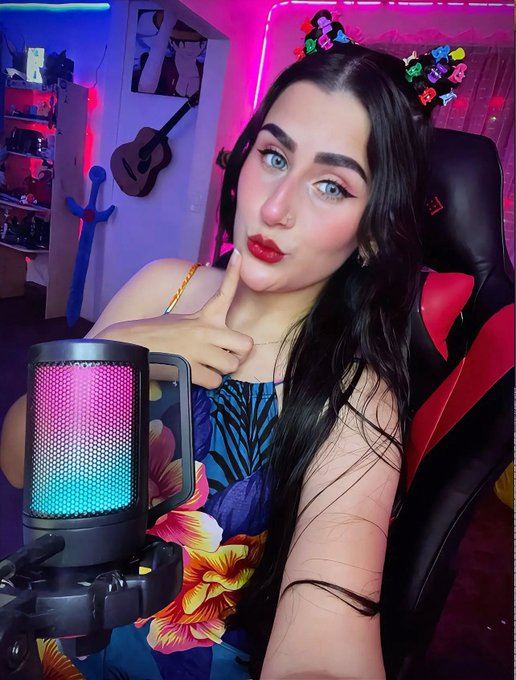 A selfie of a young woman with dark hair and a floral crown, wearing a colorful floral top, sitting in front of a rainbow LED microphone with a gaming setup visible in the background.