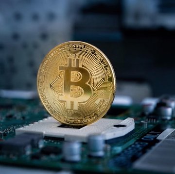 Close-up photograph of a gold Bitcoin physical coin resting on a computer motherboard with capacitors and circuit components visible.