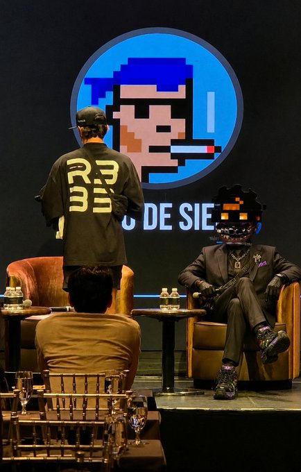 A stage event with two figures: one standing wearing a jacket that says "DREAM," and another seated wearing a suit and a pixelated black mask, with a large pixelated character projected on a circular screen behind them above partial text "DE SIE."