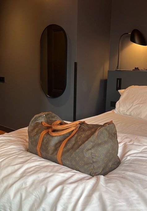 A Louis Vuitton monogram duffle bag resting on a white bed sheet in a dimly lit bedroom with a black oval mirror and lamp.