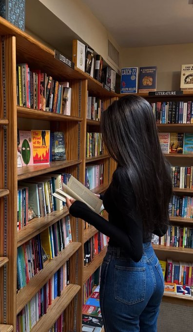 A woman with long dark hair browses bookshelves in a cozy bookstore, reading a book she pulled from the shelf, with visible section signs for "LITERATURE" and "LATIN CLASSICS." The image reflects the post's message about dedicating 30 minutes a day to learning—much like how she is taking time to read and absorb new information, the post encourages daily habits of learning, building connections, and consistently engaging on X to grow in the crypto space.