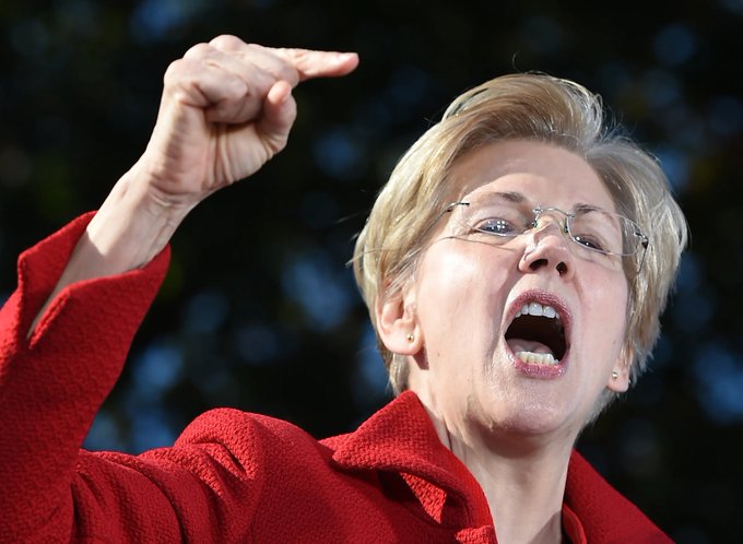 A close-up photograph of Elizabeth Warren in a red jacket, speaking animatedly with her mouth open and pointing her finger emphatically against a dark background.