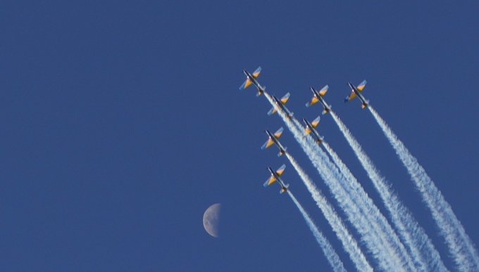 A formation of military aircraft jets flying in a V-formation leaving white smoke trails across a deep blue sky with a crescent moon visible.