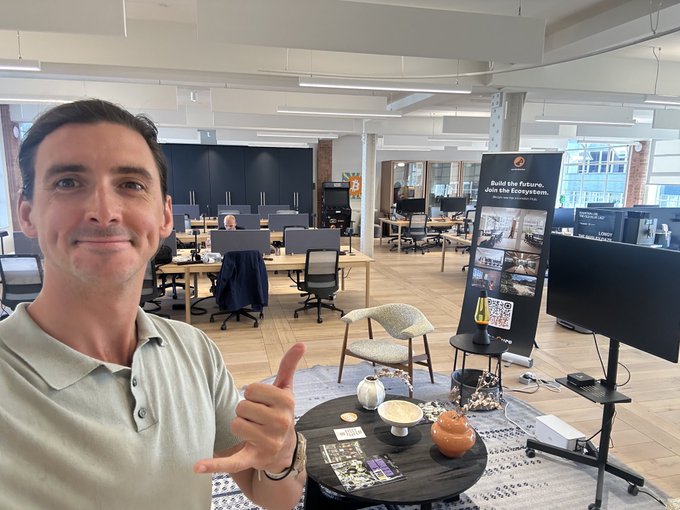 A man in a light-colored polo shirt gives a thumbs-up in an open, modern office space with desks and a recruitment banner visible in the background.