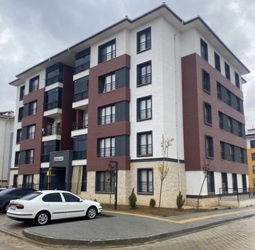 The image shows a modern four-story apartment building with a white and dark brown facade, parked cars in front, and a paved sidewalk. It contrasts with the post's commentary about buying culture—when prices are low, nobody talks about it or buys, but when prices rise, everyone discusses it and raises targets, only to end up buying at the peak, much like how property investments in such buildings often occur after prices have already surged.