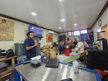 A photo of the interior of a coffee shop or roastery with several workers, a large coffee roaster, scales, and burlap coffee sacks visible.