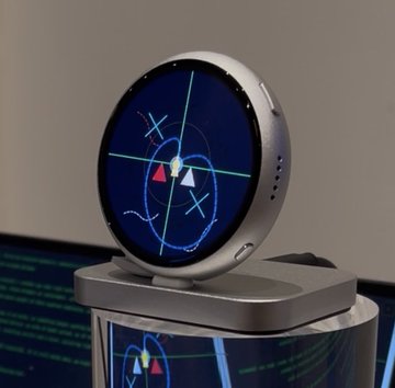 A round smartwatch displaying a dark blue watch face with a sports/play strategy design sits on a stand with a blurred laptop in the background.