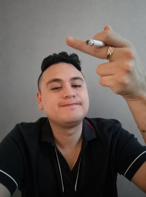 A man with a slight smile holds up his middle finger toward the camera while pinching a cigarette between his fingers, set against a plain gray background. The gesture aligns with his post's "fingerprint to the haters" message, expressing bold defiance toward critics as he shares an optimistic outlook that crypto is about to explode.