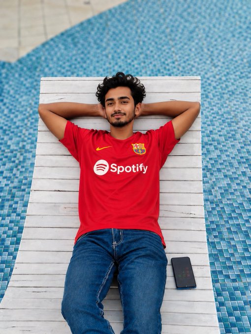 A young man wearing a red FC Barcelona jersey with 'Spotify' printed on the chest lies relaxed on a wooden lounge chair beside a blue-tiled pool.
