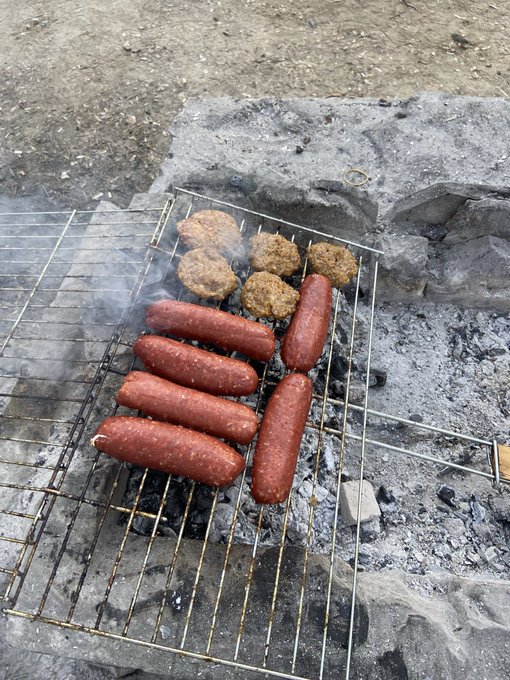 Sausages and patties sizzle over a makeshift grill of coals, with smoke curling up as the "soup makers" celebrate a successful sale with a hearty barbecue feast.