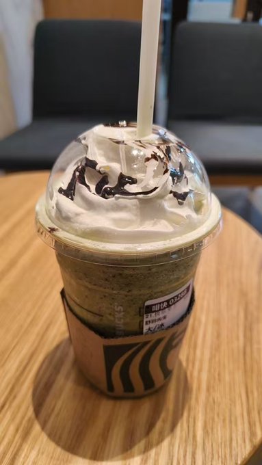 A green blended drink topped with whipped cream, chocolate drizzle, and a straw sits on a wooden table, served in a cup wrapped with a cardboard sleeve showing the "米娅の甜" branding. The image complements the post's message of finding comfort and healing in small daily pleasures—like treating yourself to a sweet pick-me-up drink during slower market days.