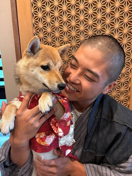 A smiling young man in a gray kimono holds a Shiba Inu dressed in a red floral kimono, both appearing joyful in the warm embrace. The image's tender moment between owner and pet reflects the post's tone of affection and reconciliation with the $SAN team, suggesting warmth and goodwill despite past disagreements.