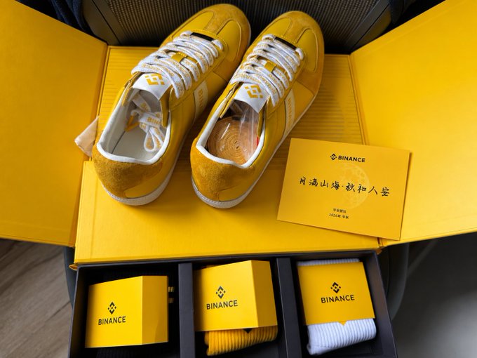 A pair of yellow sneakers with the Binance logo sits in branded Binance packaging, accompanied by socks, a card with Chinese calligraphy reading '月满山辉秋扣人堂', and additional branded boxes.
