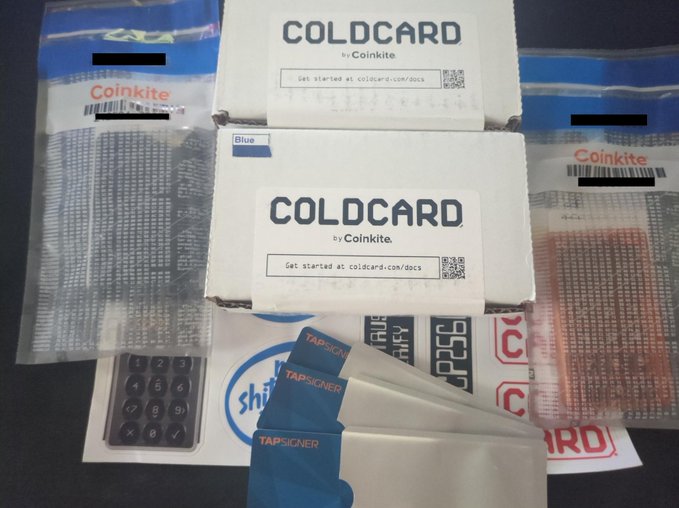 Two unopened Coldcard by Coinkite boxes (including a Blue Mk5) sit alongside packaged TAPSIGNER cards, Clear Case accessories, and Coinkite-branded stickers and component bags, showcasing the hardware wallet merchandise that was received to support the Lab312 and MarseilleBTC meetups.