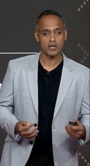 A man in a light blue blazer and black shirt speaking on stage while gesturing with both hands.