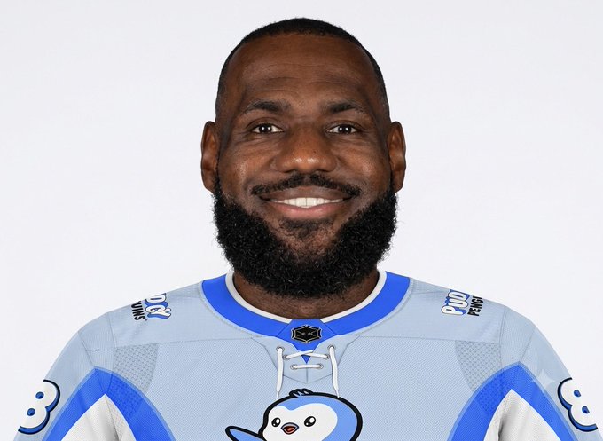 A headshot of NBA player LeBron James smiling, wearing a light blue and white jersey with a penguin logo.