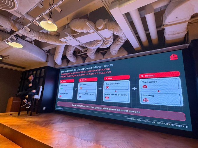 A person sits on a stool presenting a slide titled 'Illustrative Multi-Asset Cross-Margin Trade' to a room with exposed ceiling pipes, describing how unified collateral can unlock use cases like trading, using services, and investing in treasuries.