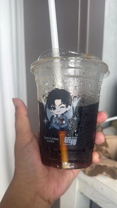 A hand holds an iced coffee drink in a clear plastic cup branded with a Cotti Coffee (库迪咖啡) cartoon logo featuring a boy in a hoodie.