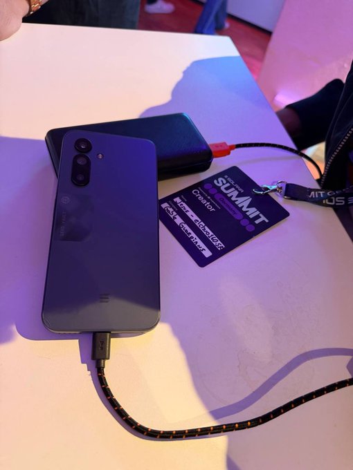 The image shows an old cell phone connected to a charger with a black cord. The phone has a purple screen that displays "I spent the whole day at the @solana Summit Berlin event hosted by @SuperteamDE" written on its back, indicating that someone was there and took photos or recorded videos during their time at the event.