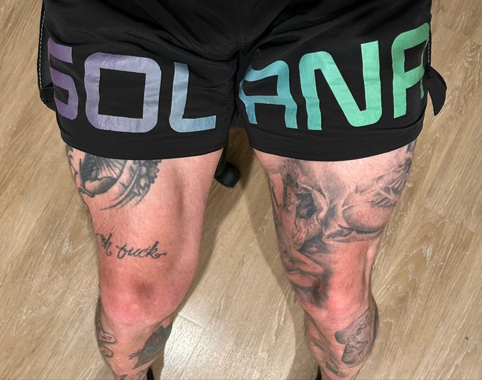 A view of a person's tattooed legs wearing black MMA shorts with 'SOL ANF' written across the front in gradient colors, with skull and 'oh, ducks' text tattoos visible on the thighs.