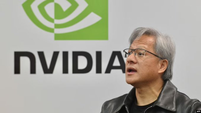 The image features a man wearing glasses, standing in front of a large green sign that says "NVDA" with an arrow pointing towards him. The man appears to be speaking or giving a presentation, as he is positioned behind the sign. The background of the image is white, which contrasts with the green sign and makes it stand out more prominently.