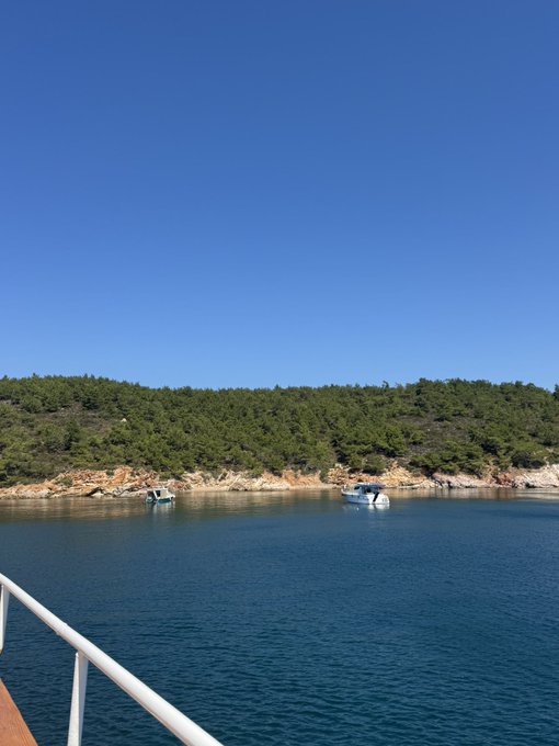 A scenic photograph taken from a boat railing shows two small boats anchored in clear blue water near a rocky, forested coastline under a bright blue sky.