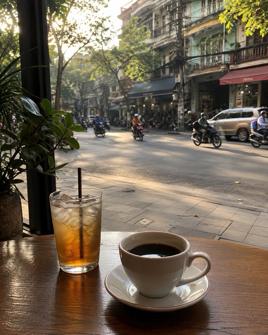 A coffee and iced tea sit on a wooden café table by the window, overlooking a Hanoi's street scene with motorbikes and pedestrians passing by in the morning light.

The image perfectly captures the cozy atmosphere described in the post—a peaceful café moment with hot coffee in hand, watching the city slowly come alive as morning traffic builds up.