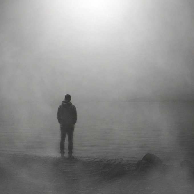 A grayscale, atmospheric image shows a solitary figure in a hoodie standing at the edge of a misty body of water.