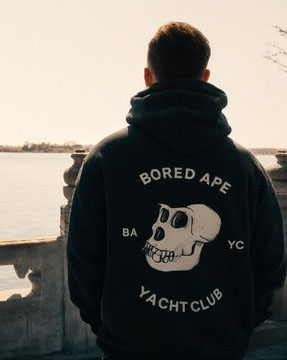 A person stands by a body of water wearing a black 'BORED APE YACHT CLUB' hoodie featuring the iconic skull logo and 'BAYC' lettering.