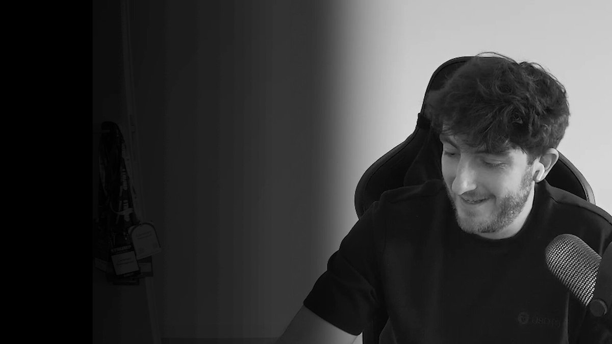 The image features a man sitting at a desk with headphones on, wearing a black shirt. The background appears to be a plain white wall or gray surface. There are no texts or other objects visible in the image.