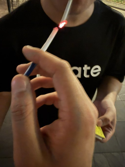 A person in a black "Gate" t-shirt holds a glowing stick, lighting the tip with a flame while another hand reaches in from the foreground, matching the post's playful clue about "Brothers who landed without fire" looking for "Brother Lu" to ignite "2049."