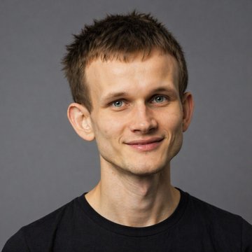 A portrait of a smiling man with short spiky brown hair and blue eyes, wearing a black t-shirt against a gray background.