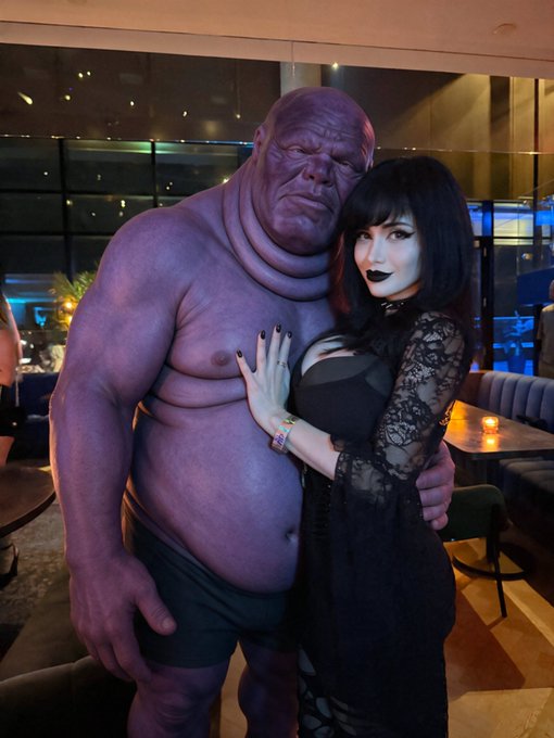 A woman with dark hair, dark lipstick, and a black lace dress poses next to a person in a full-body purple Thanos cosplay costume.