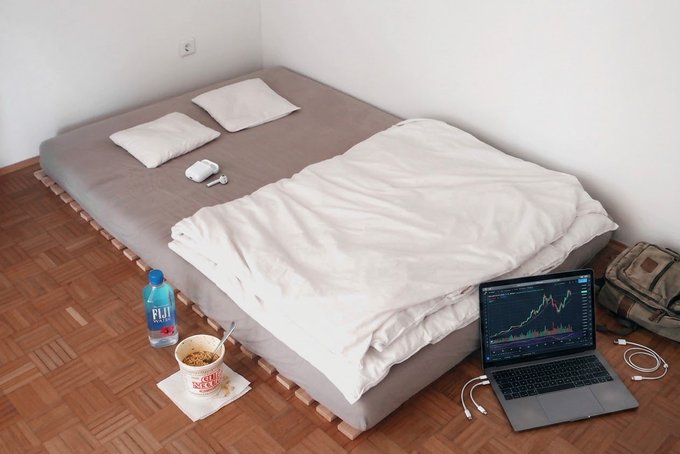 A minimalist bedroom with a low mattress on a wooden pallet, accompanied by a Fiji water bottle, a cup of Cup Noodles, and a laptop displaying a stock chart.