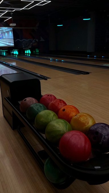 A bowling alley scene shows a ball return rack loaded with colorful bowling balls—red, green, orange, and purple—leading down wooden lanes lit by neon pin lights.