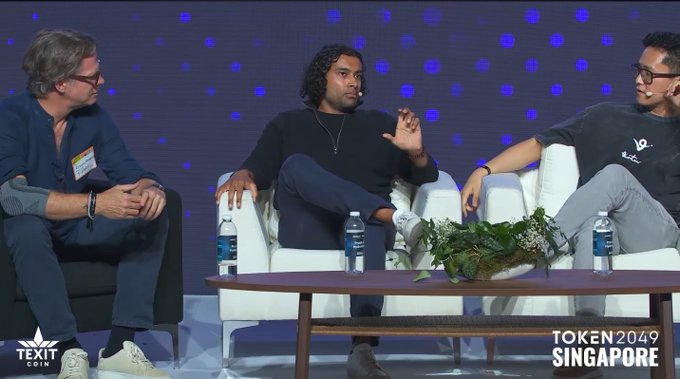 Three men sit on white chairs on a stage at the TOKEN2049 Singapore conference, with one man in the center speaking while gesturing.
