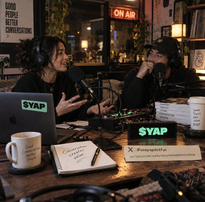Two people wearing headphones are hosting a podcast in a studio with an 'ON AIR' sign visible; a laptop displays a '$YAP' sticker and a sign reads '$YAP' with the handle '@useyapdotfun'.