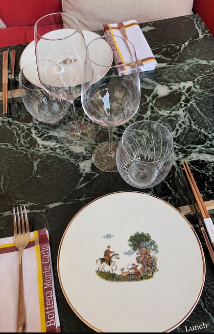 The image features a dining table with various items arranged on top of it. There are two wine glasses placed next to each other, along with a plate that has a picture or painting depicting horses on it. The table also includes forks, knives, and spoons, suggesting that this is set up for a meal. The background appears to be black, which contrasts with the white plates and glassware. The image seems to be related to an advertisement promoting a lifestyle centered around art and culture.