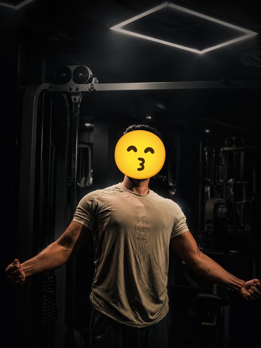 A muscular man in a beige t-shirt stands in a dimly lit gym with a kissing-face emoji covering his face.