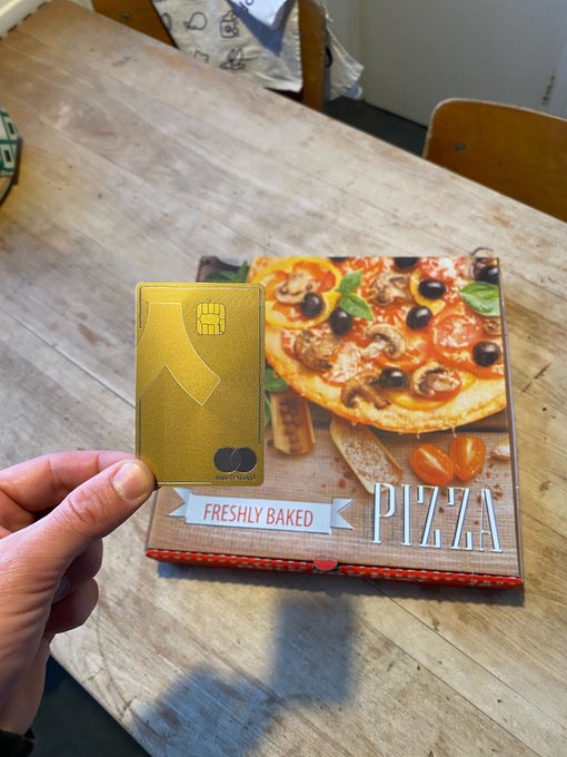 The image shows a person holding up a pizza box that has a pizza inside, along with an American Express card. The card appears to be a Mastercard or similar payment method. This suggests that the person might have used their credit card for purchasing the pizza.