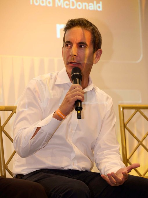 A man with short brown hair wearing a white button-up shirt speaks into a microphone while seated, with partial text 'Todd McDonald' visible on the projection screen behind him.