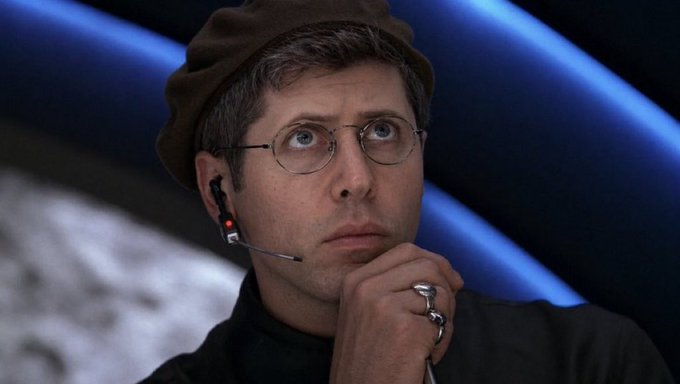 A man wearing a brown beret, round glasses, and a headset microphone, resting his chin on his hand with a thoughtful expression.